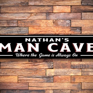 May include: Black and white sign with the text "NATHAN'S MAN CAVE Where the Game is Always On". The sign has a red border and is mounted on a wood wall.