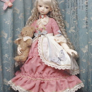 Op de afbeelding: Een porseleinen pop die een roze jurk draagt met witte kant en een kroon. De pop houdt een teddybeer vast en heeft blond haar. De achtergrond is een blauw en wit bloemenpatroon.