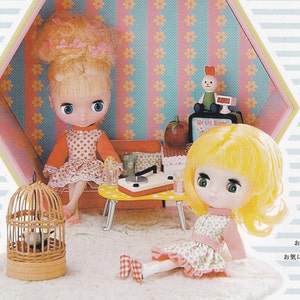 May include: Two dolls with blonde hair and large eyes are sitting in a dollhouse. The dollhouse has pink and blue striped wallpaper with yellow flowers. One doll is wearing a red and white polka dot dress and the other is wearing a green and white floral dress. The dollhouse also has a small table with a record player and a birdcage.