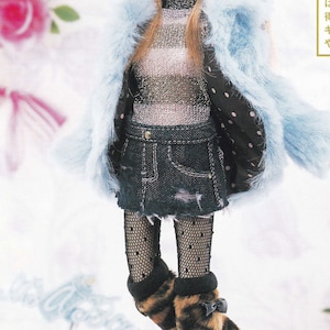 May include: A doll with long brown hair wearing a blue faux fur coat, a silver and black striped shirt, a denim skirt, black fishnet tights, and leopard print boots. The doll is standing on a white surface with a pink background. The text "かな フリル も いい か も ね" is written in Japanese on the background.
