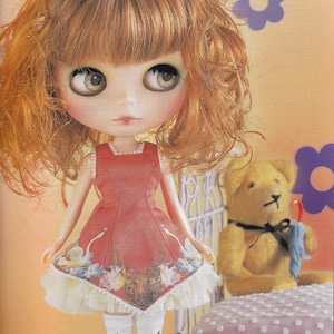May include: A doll with long, curly, brown hair wearing a red dress with a white lace trim and white tights with a floral pattern. The doll is standing in front of a white metal crib with a pink and white polka dot mattress.