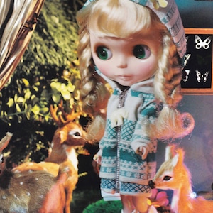 May include: A doll with blonde hair wearing a green and white hooded sweater with a white trim. The doll is standing in front of a green leafy background with two deer figurines.