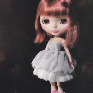 May include: A doll with long, red hair wearing a light gray tulle dress with a white lace trim. The doll has pink eyeshadow and pink lips.