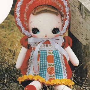 Könnte beinhalten: Eine handgemachte Puppe mit einer rot-weißen Haube, einem roten Kleid mit weißer Spitzenborte und einem blau-weiß gemusterten Rock mit roten Äpfeln. Die Puppe sitzt auf einem Strohboden.