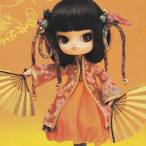 May include: A doll with black hair and large, blue eyes is wearing a pink and orange kimono with gold accents. The doll is holding two gold fans. The background is a yellow and gold pattern.