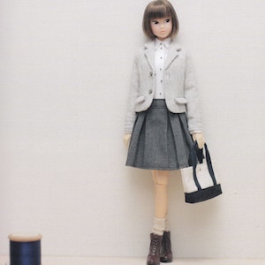 Puede incluir: Una muñeca con un blazer gris, una falda plisada gris, una camisa blanca y botas marrones. La muñeca lleva un bolso de mano blanco y negro.