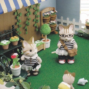 Puede incluir: Un conjunto de juguetes en miniatura con tres gatos de pelaje marrón y blanco. Los gatos llevan ropa y están de pie sobre una superficie verde cubierta de hierba. El conjunto incluye una pequeña valla blanca de madera, un jardín en miniatura con flores y un pequeño cartel de madera que dice "Happy Garden".