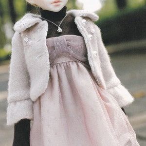 May include: A doll wearing a white knitted jacket with pearl accents, a pink tulle dress with a bow, and black mock neck shirt. The doll has blonde hair and blue eyes. The doll is standing on a sidewalk with green foliage in the background. The text "Finding somethin new" is written on a piece of paper in the upper right corner of the image.