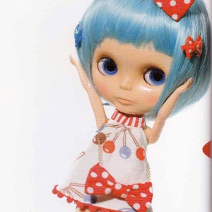 May include: A doll with blue hair and a red and white polka dot bow. She is wearing a white dress with red and blue polka dots and a red bow. The dress has a white trim with red pom-poms.