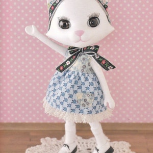 Puede incluir: Una muñeca de gato blanco con un vestido azul y blanco con estampado floral y una diadema negra con estampado floral. La muñeca está de pie sobre un encaje de ganchillo blanco.