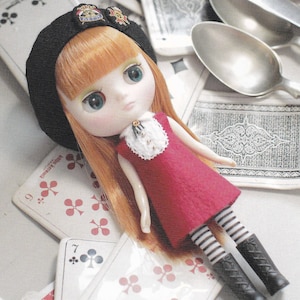 May include: A doll with red hair wearing a black beret, a red dress with white lace trim, and black and white striped tights. The doll is lying on a table with playing cards.
