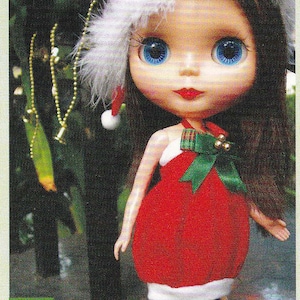 May include: A doll wearing a red and white dress with a green bow and a white fur trimmed hat. The doll has blue eyes and long brown hair. The doll is standing in front of a green background with a string of beads hanging from the top.