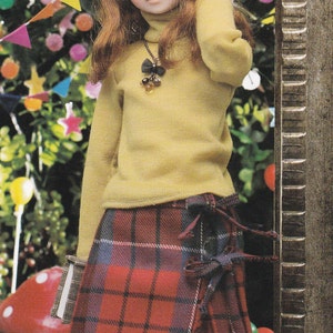 May include: A doll wearing a yellow sweater with a black and gold necklace and a red and black plaid skirt.