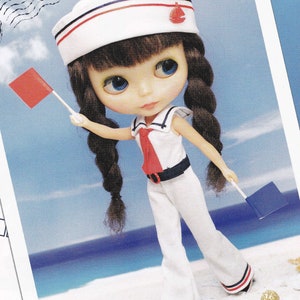 May include: A doll dressed in a sailor outfit, holding two small flags. The doll has brown braided hair, blue eyes, and is wearing a white sailor hat with red and blue stripes. The outfit is white with red and navy accents.