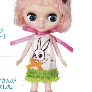 Puede incluir: Una muñeca con el pelo rosa, ojos azules y un vestido blanco con un diseño de conejo negro. La muñeca lleva zapatos verdes y blancos.