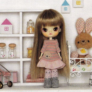 May include: A doll with long brown hair wearing a pink and grey striped dress with a bear applique. The doll has blue eyes and is standing in front of a white shelf with jars and a small bed with a brown bunny doll.