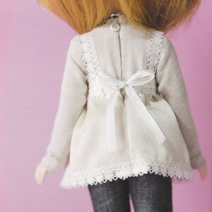 May include: A white doll dress with lace trim and a white ribbon tied at the back. The dress has a long sleeve and a slightly gathered skirt.