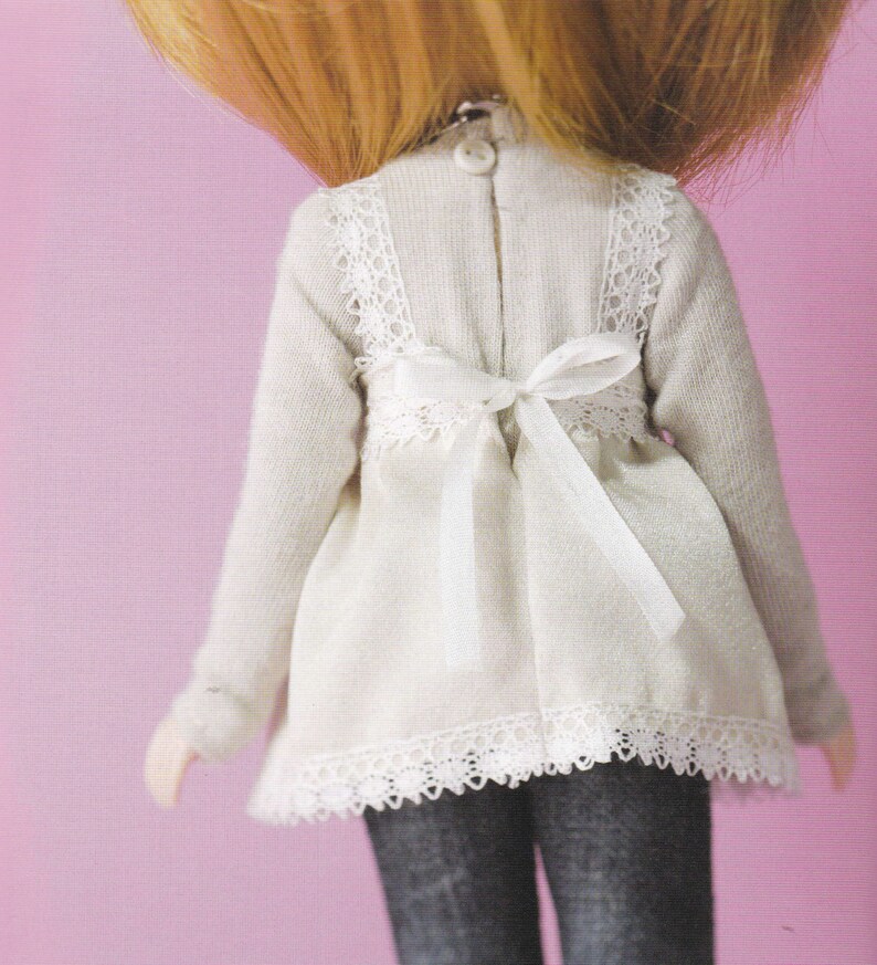 May include: A white doll dress with lace trim and a white ribbon tied at the back. The dress has a long sleeve and a slightly gathered skirt.