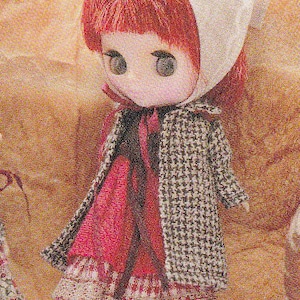 Puede incluir: Una muñeca con un vestido rojo y blanco, un abrigo de estampado marrón y blanco y un gorro blanco con bordados rojos. La muñeca tiene el pelo rojo y grandes ojos azules.