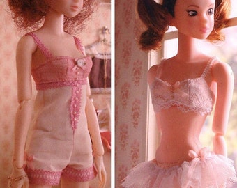 May include: Two dolls wearing pink lingerie. The doll on the left is wearing a pink and white lace one-piece with a floral embellishment. The doll on the right is wearing a pink and white lace bra and a tulle skirt.
