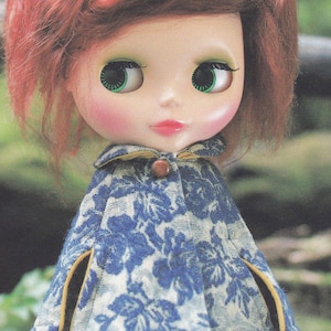 May include: A doll with long, red hair wearing a blue and white floral print cape with yellow trim. The doll has green eyes and rosy cheeks.