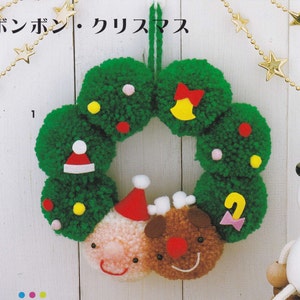 以下が含まれることがあります： 赤いリボン、黄色のベル、ピンクのリボン、赤いサンタの帽子、茶色のトナカイの顔で飾られた緑のポンポンリース。リースは、金の星が連なったチェーンが飾られた白い壁にかけられています。