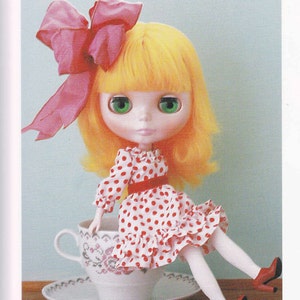 Puede incluir: Una muñeca con cabello rubio y ojos verdes lleva un vestido de lunares rojos y blancos y zapatos rojos, sentada en una taza de té blanca con un patrón floral.