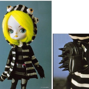 Może przedstawiać: Lalka w czarno-białe paski z żółtymi włosami, ubrana w czarno-białą kurtkę w paski z kapturem i dużymi oczami. Lalka trzyma czarną torbę z kolcami.