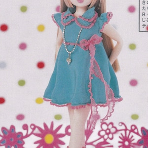 May include: A doll with long blonde hair wearing a blue dress with pink lace trim and a pink bow. She is wearing pink socks and white shoes. The background is a white surface with pink and green polka dots.