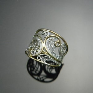 May include: A silver and gold filigree ring with a wide band. The ring is made of two intertwined bands, one silver and one gold, and features a delicate, intricate design.