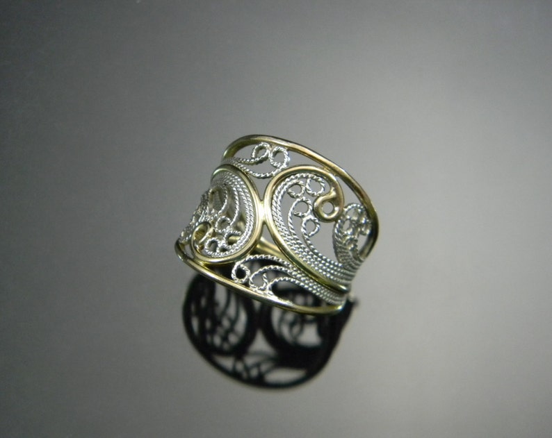 May include: A silver and gold filigree ring with a wide band. The ring is made of two intertwined bands, one silver and one gold, and features a delicate, intricate design.