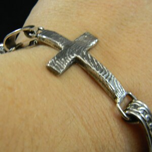 May include: A silver cross bracelet with a textured design. The bracelet is worn on a person's wrist.