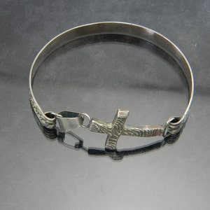 May include: A silver bracelet with a textured cross charm. The bracelet is made of a thin band of metal and has a simple design.