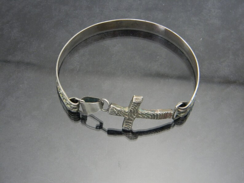 May include: A silver bracelet with a textured cross charm. The bracelet is made of a thin band of metal and has a simple design.