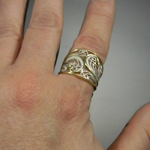 May include: A wide silver ring with a gold filigree design. The ring is worn on a finger.