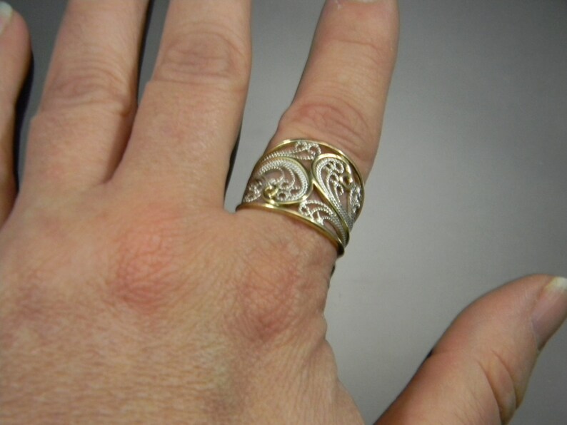 May include: A wide silver ring with a gold filigree design. The ring is worn on a finger.