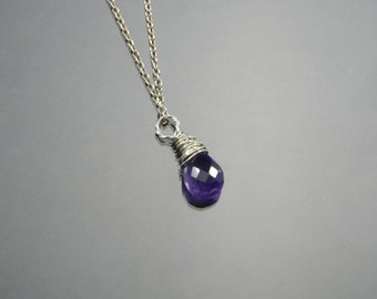 May include: A silver necklace featuring a faceted, deep purple amethyst pendant. The gemstone is wrapped in silver wire and hangs from a delicate chain. The necklace is displayed against a reflective, gray background.