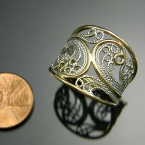 May include: A silver filigree ring with a gold band. The ring is wide and has a delicate, intricate design.
