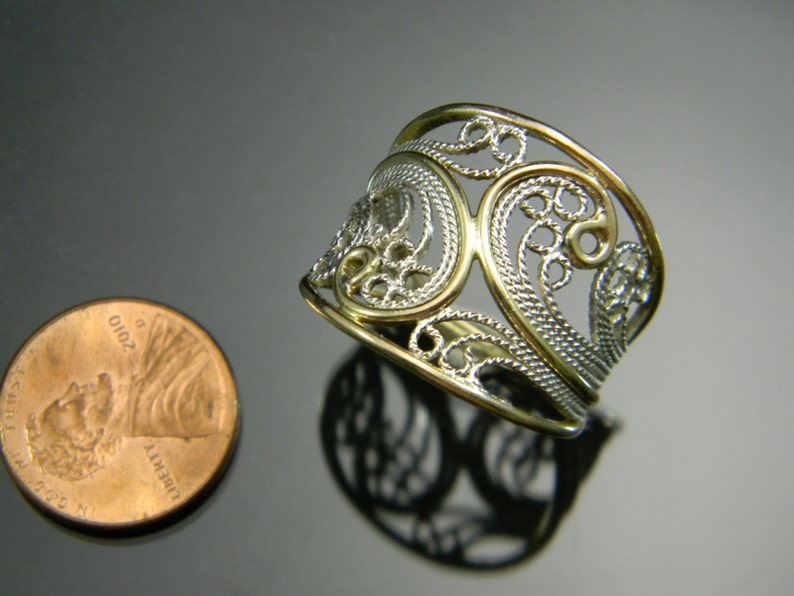 May include: A silver filigree ring with a gold band. The ring is wide and has a delicate, intricate design.