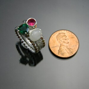 May include: A silver ring with three stones: a green stone, a pink stone, and a white stone. The ring is next to a US penny for size comparison.