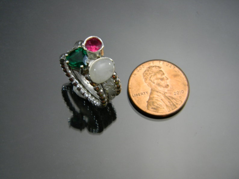 May include: A silver ring with three stones: a green stone, a pink stone, and a white stone. The ring is next to a US penny for size comparison.