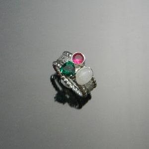 May include: A silver ring with three stones: a green heart-shaped stone, a pink round stone, and a white oval stone.