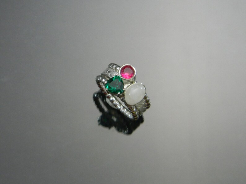 May include: A silver ring with three stones: a green heart-shaped stone, a pink round stone, and a white oval stone.