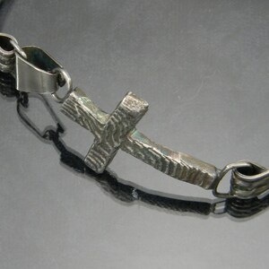May include: A silver bracelet with a textured cross charm. The bracelet is made of a thin metal band and has a simple design.