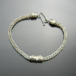 May include: A silver bracelet with a braided chain design and a decorative silver accent. The bracelet has a clasp closure.