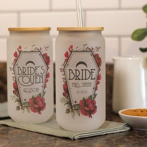 May include: Two frosted glass tumblers with gold lids and black and white graphics. The tumblers feature a floral design with red roses and the text "Bride's Coven" and "Bride".