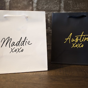 Pode incluir: Duas sacolas de presente brancas com al&ccedil;as pretas. Uma sacola tem o nome "Maddie xoxo" impresso em preto. A outra sacola tem o nome "Austin xoxo" impresso em dourado.