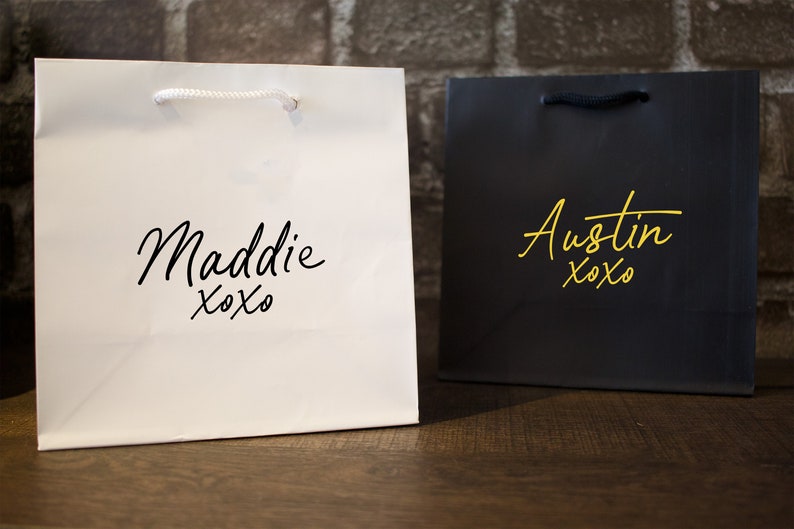 Pode incluir: Duas sacolas de presente brancas com al&ccedil;as pretas. Uma sacola tem o nome "Maddie xoxo" impresso em preto. A outra sacola tem o nome "Austin xoxo" impresso em dourado.