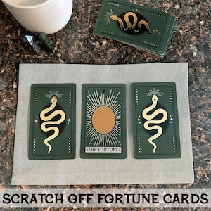 May include: Three green fortune cards with gold foil accents. The cards feature a snake, a sun, and a snake. The text on the middle card reads "The Fortune".