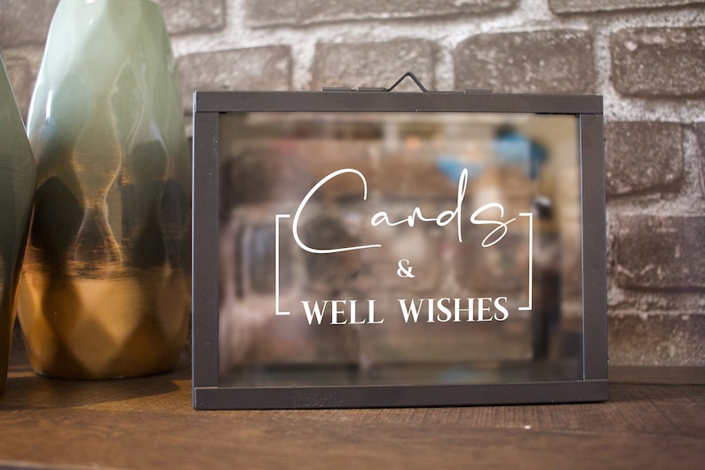May include: A rectangular, dark gray metal card box with a glass front. White cursive text on the glass reads "Cards & Well Wishes". Two decorative vases are visible to the left of the box. The background is a brick wall.